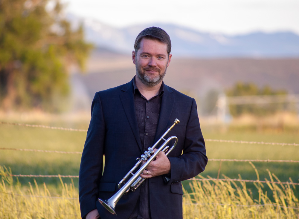 Eddie holding a trumpet outdoors with blurred mountains in the background